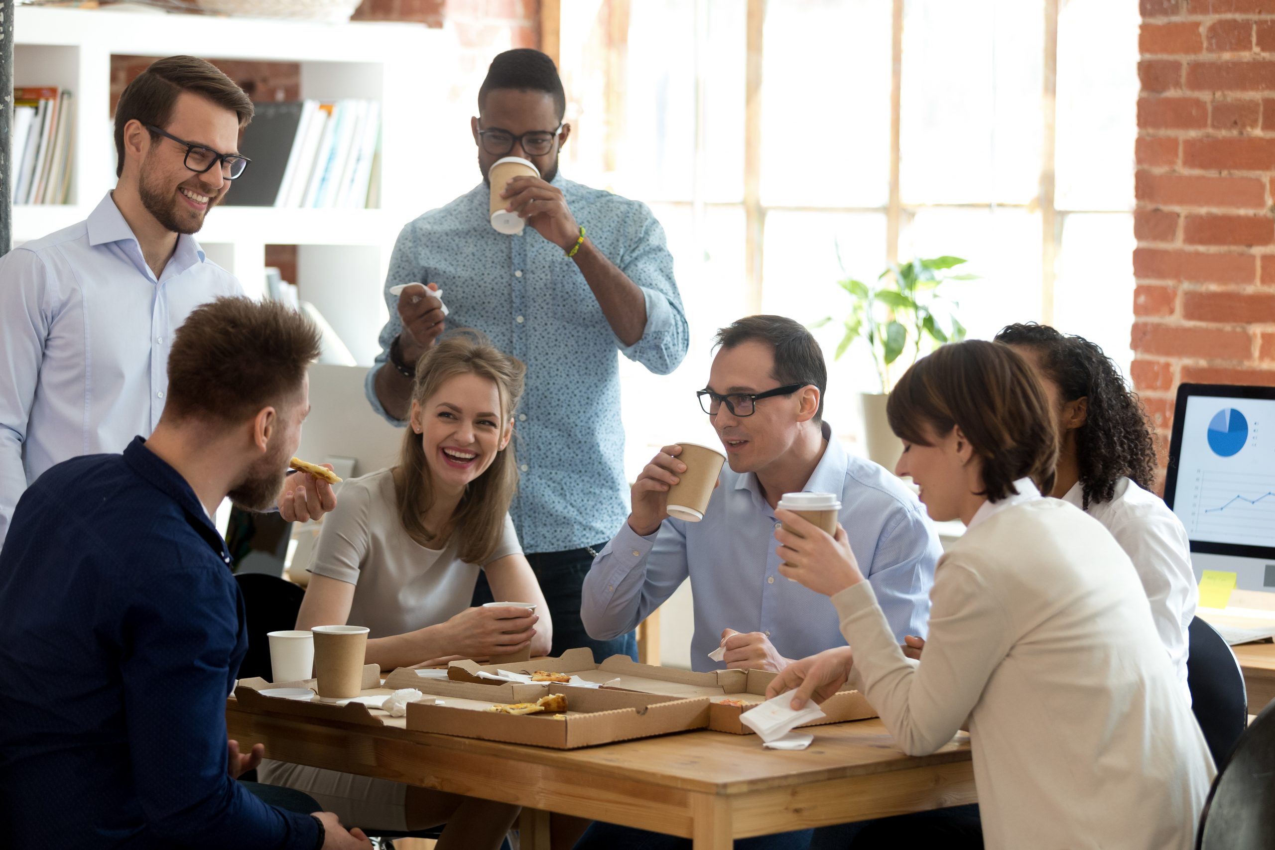 Happy diverse team talking having fun drinking coffee eating pizza together, multi-ethnic employees group enjoy takeaway food friendly conversation share lunch chatting laughing at office work break